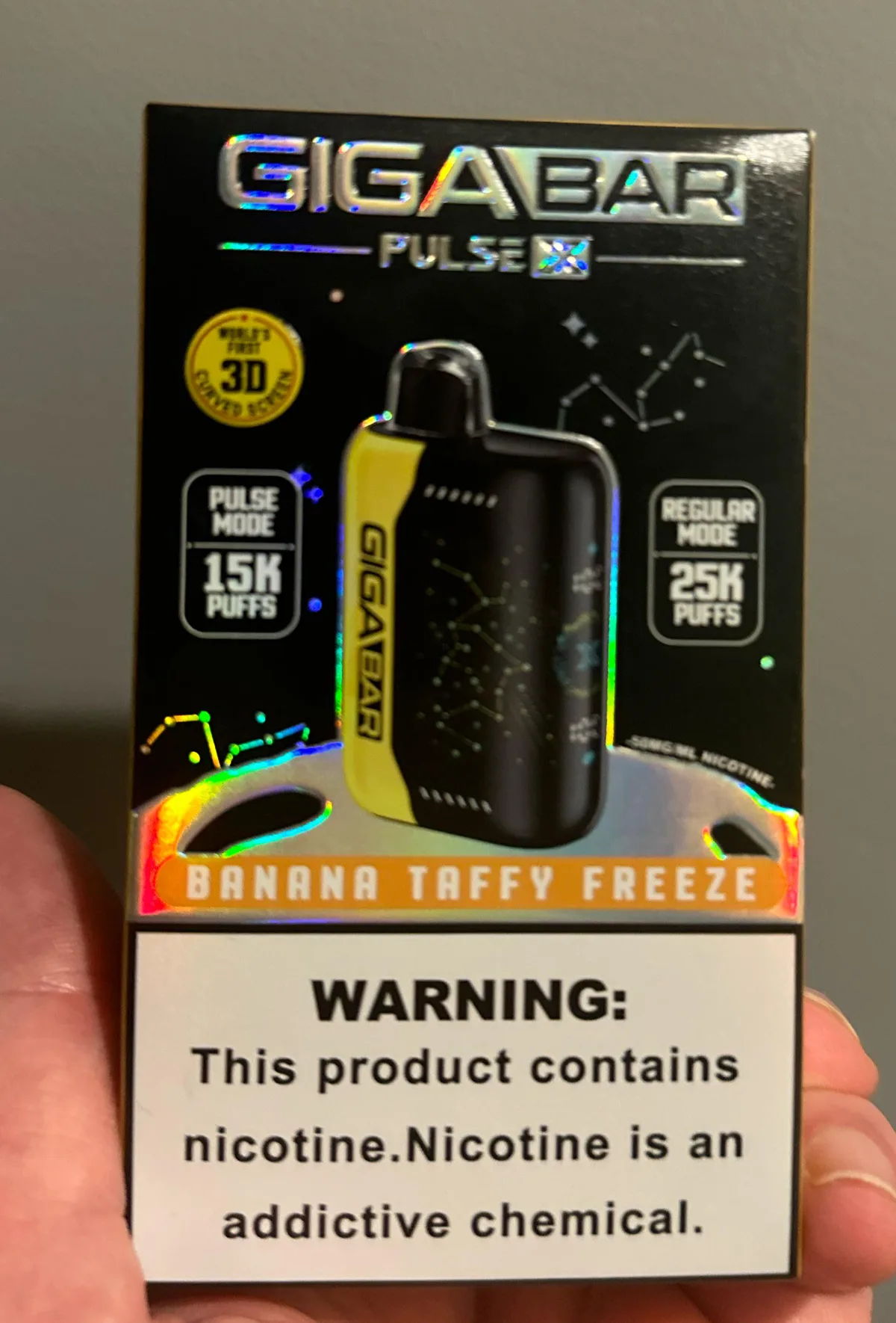 A hand holding a boxed disposable vape branded GIGABAR PULSE X, with a badge reading World's First 3D Curved Screen, Pulse Mode 15K puffs and Regular Mode 25K puffs, flavor Banana Taffy Freeze, and a nicotine warning on the bottom third of the box.