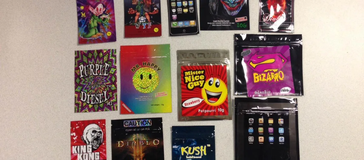 Seized foil packets laid on a table: Purple Diesel, Mr. Happy, Mister Nice Guy Strawberry Potpourri 10g, Bizarro, King Kong, Diablo, Kush, and packets printed to look like a smartphone.