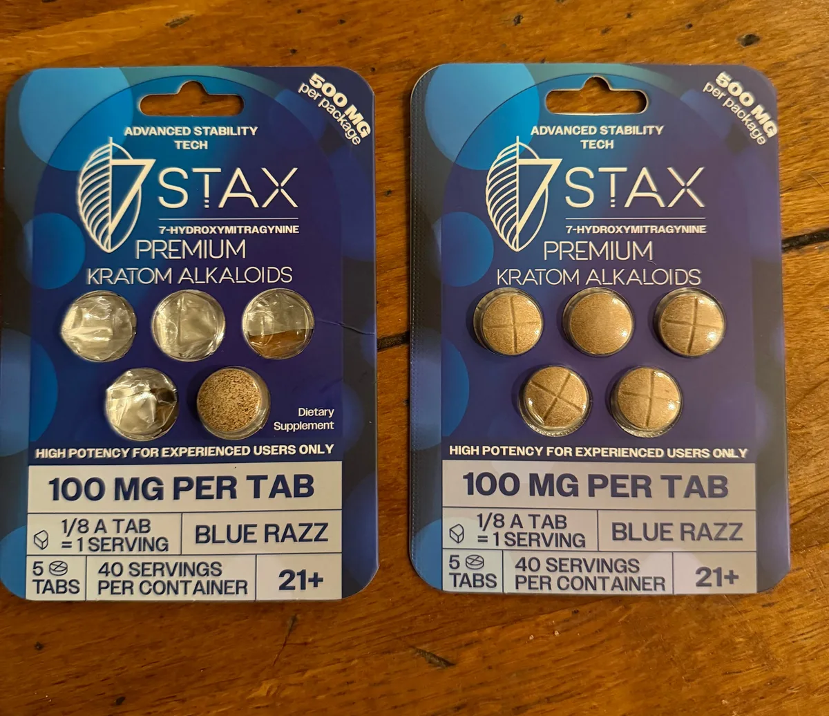 Two blue 7 Stax blister cards side by side on a wooden table, both 7-Hydroxymitragynine Premium Kratom Alkaloids, 100 MG PER TAB, Blue Razz. The left card is opened and printed Dietary Supplement; the right card is sealed and has no such line.