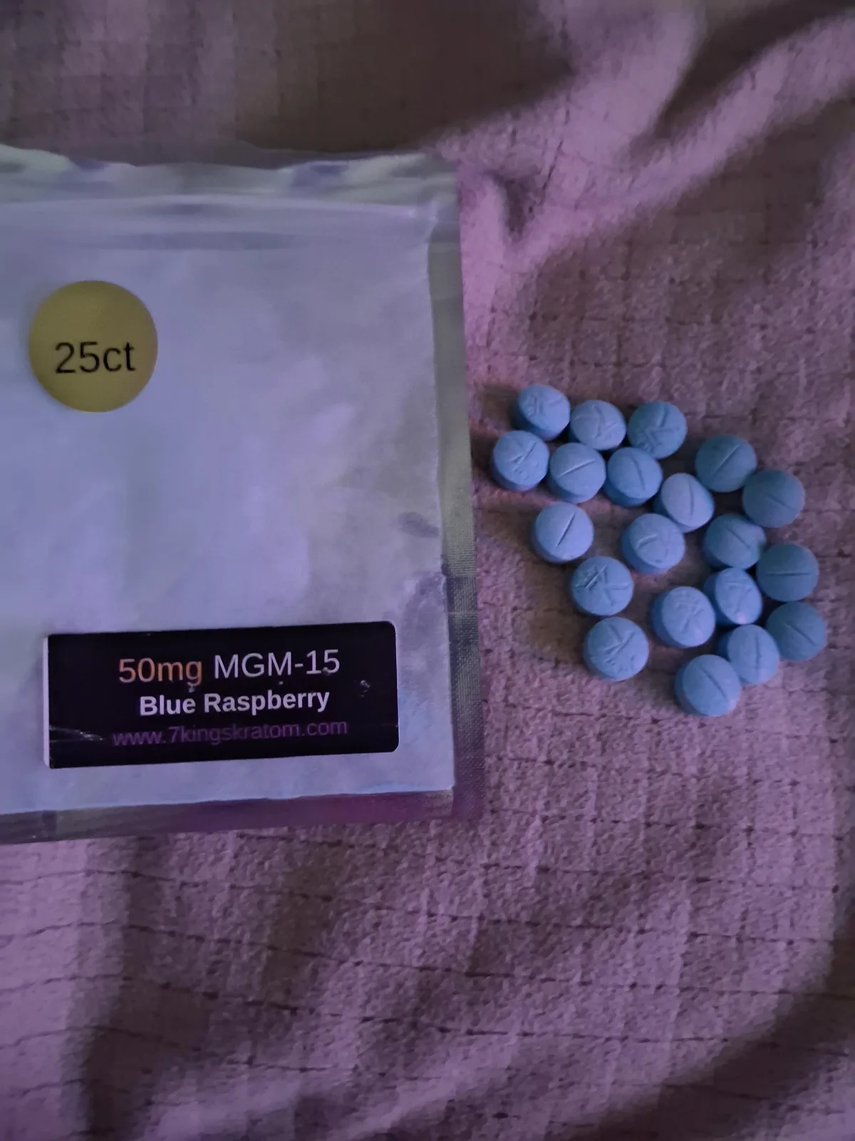 A foil bag with a 25ct sticker and a label reading 50mg MGM-15 Blue Raspberry, www.7kingskratom.com, beside twenty blue scored tablets on a sheet.