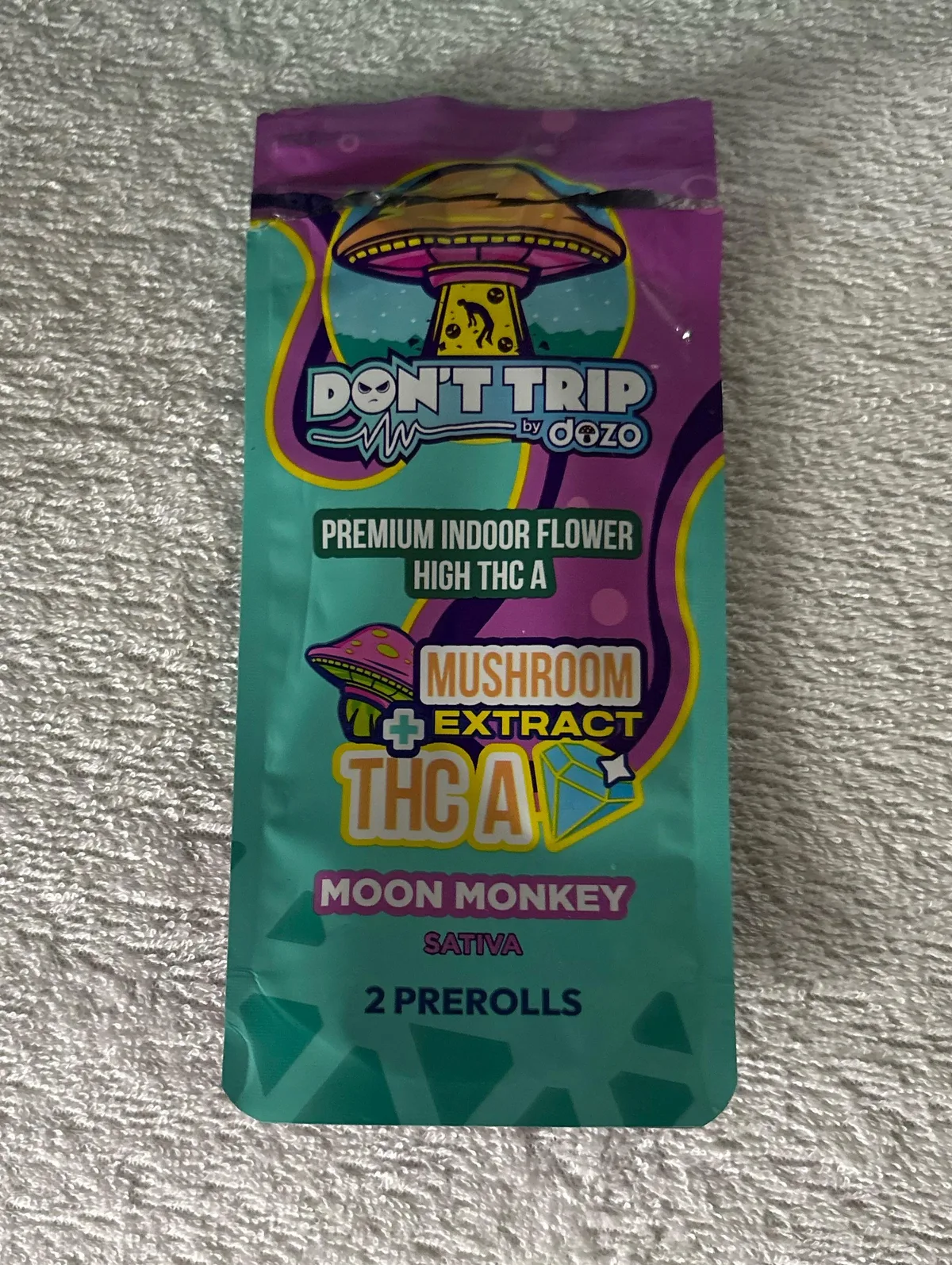A teal and purple pouch reading Don't Trip by dozo, Premium Indoor Flower High THC A, Mushroom Extract plus THC A, Moon Monkey Sativa, 2 prerolls, with a flying saucer drawn above.