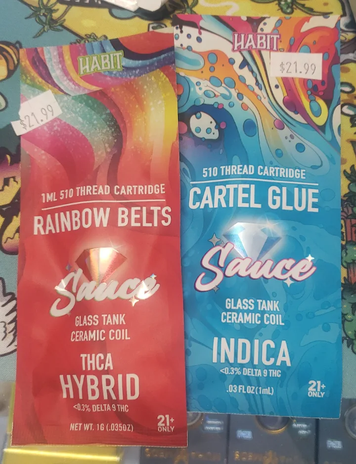 Two Habit Sauce cartridge sleeves on a smoke-shop counter, Rainbow Belts and Cartel Glue, each with a 21.99 dollar price sticker, marked 510 thread cartridge, glass tank, ceramic coil, under 0.3% delta 9 THC, 21+ only.