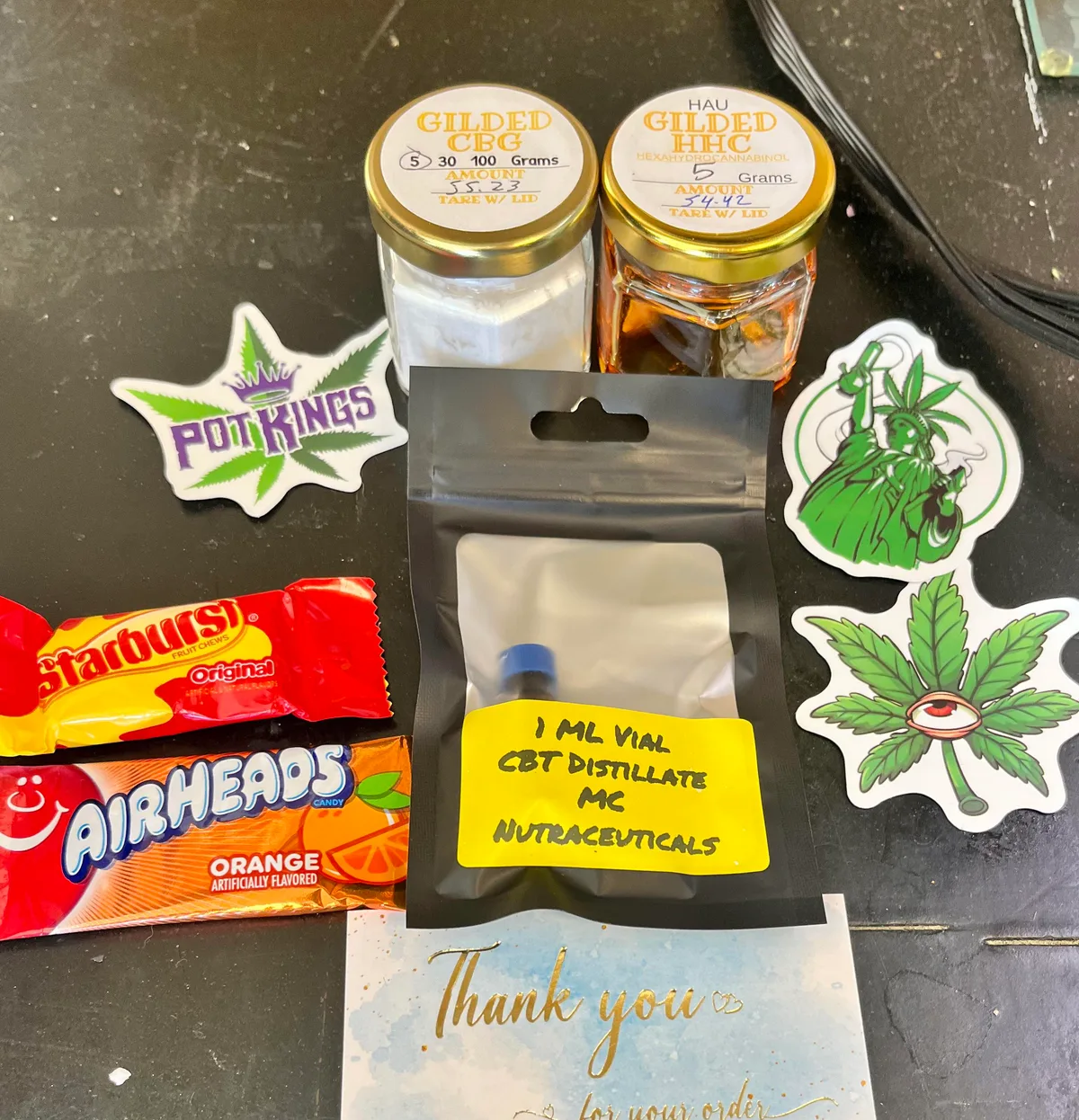 A buyer's photo of a Gilded Extracts order: a jar of CBG and a jar labelled Gilded HHC with HAU written above, a small bag holding a 1 ml vial of CBT distillate labelled MC Nutraceuticals, stickers, candy and a thank-you card.