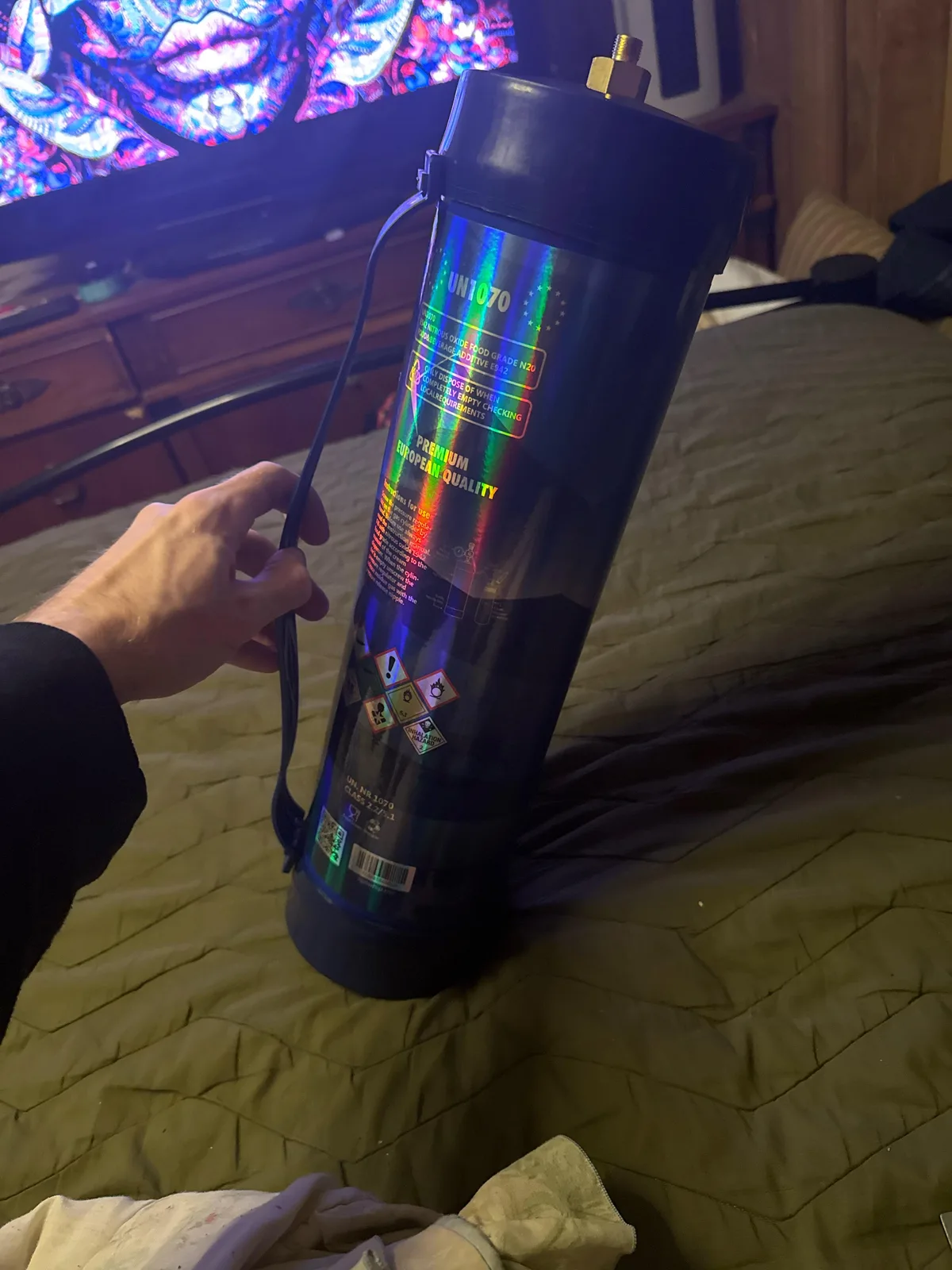 A hand holding a dark holographic nitrous tank by a black strap, on a bed. The label reads UN1070, Nitrous Oxide Food Grade N2O, Premium European Quality, and shows hazard symbols including Inhalation Hazard.