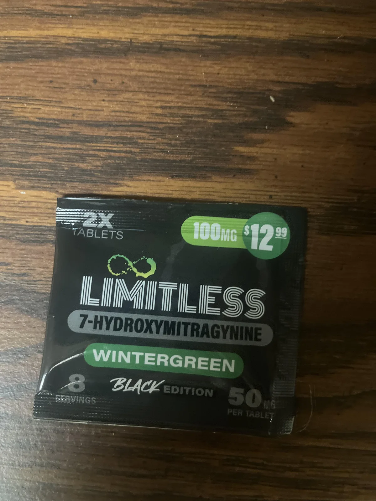 A black foil pouch on a wooden table: 2X tablets, 100 MG, 12.99 dollars, LIMITLESS 7-HYDROXYMITRAGYNINE, Wintergreen, Black Edition, 8 servings, 50 mg per tablet.