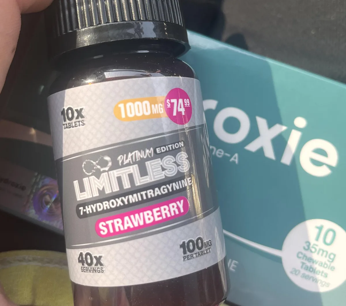A hand holding a black bottle with a silver label: 10X tablets, 1000 MG, 74.99 dollars, Platinum Edition, LIMITLESS 7-HYDROXYMITRAGYNINE, Strawberry, 40X servings, 100 mg per tablet, beside another brand's box.