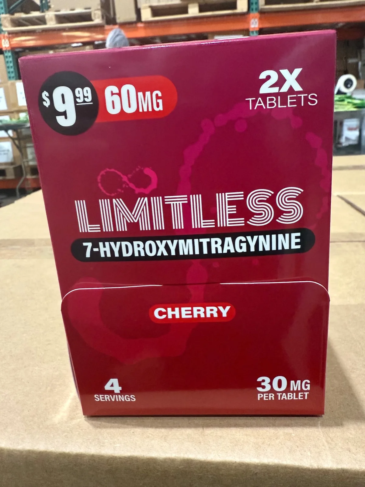 A red display box on cartons in a warehouse: 2X tablets, a printed price of 9.99 dollars for 60 mg, the word LIMITLESS in striped letters under an infinity symbol, 7-HYDROXYMITRAGYNINE, Cherry, 4 servings, 30 mg per tablet.