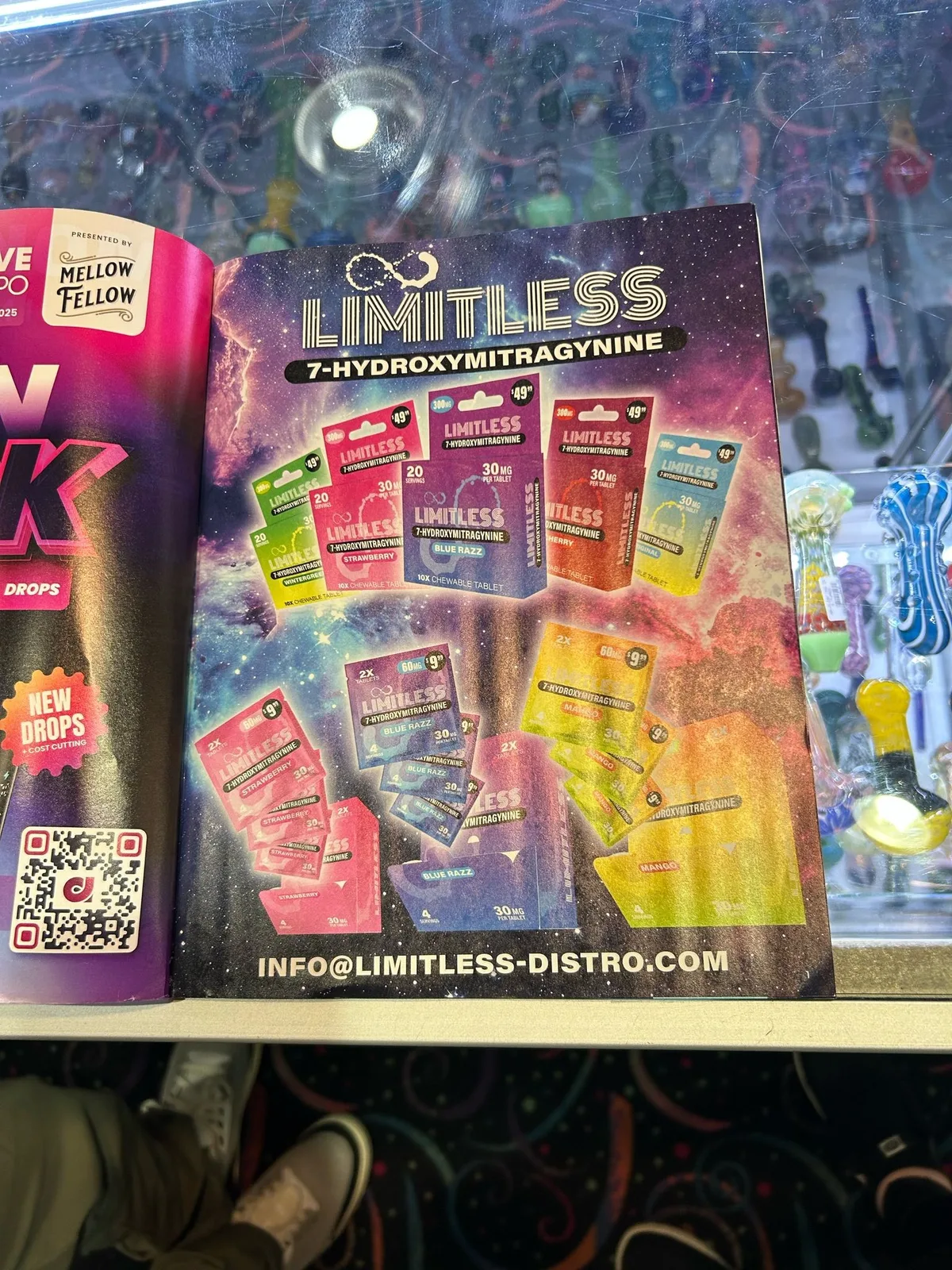 A magazine lying open on a glass display case of pipes: a full-page advertisement headed LIMITLESS 7-HYDROXYMITRAGYNINE showing 10-count boxes in five flavours priced 49 dollars and two-tablet packs priced 9.99, with the line INFO@LIMITLESS-DISTRO.COM at the foot.
