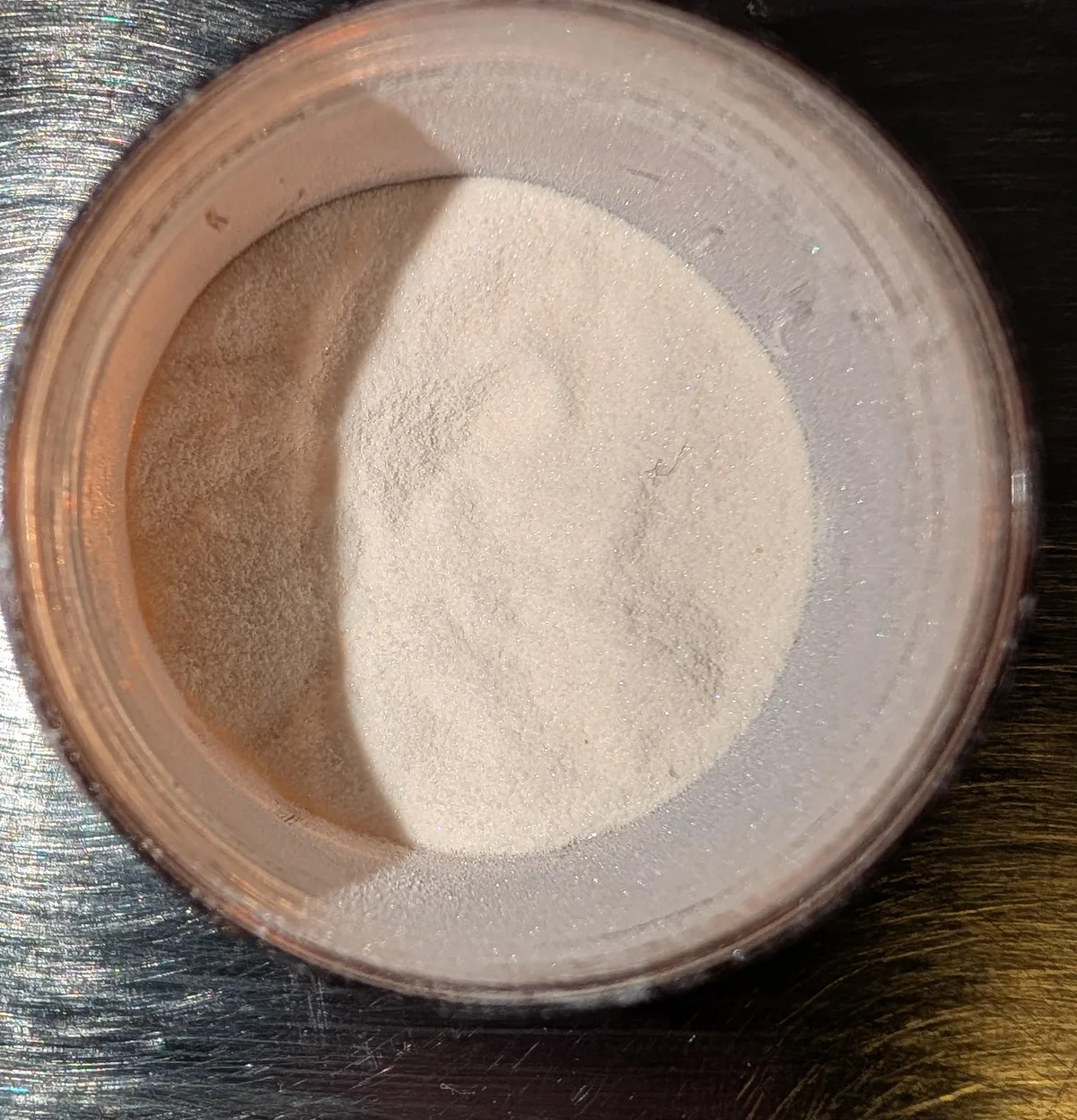 A photograph looking down into a small clear jar of fine off-white powder with a faint orange tint, on a steel surface.