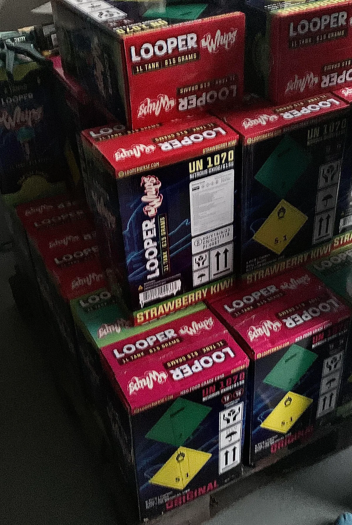 A buyer's photograph of stacked red and black cartons printed LOOPER Whips, 1L tank, 615 grams, in Strawberry Kiwi and Original, with UN 1070 nitrous oxide markings and hazard diamonds.