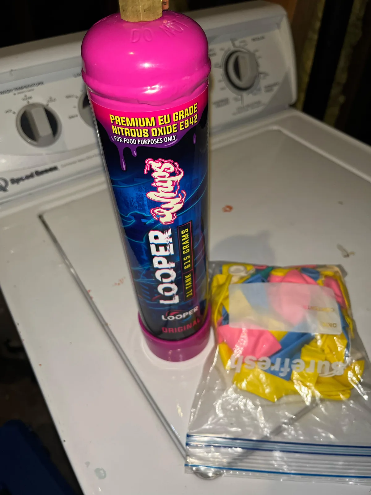 A pink-topped Looper Whips tank, 1L, 615 grams, Original, beside a bag of balloons on a washing machine. The label reads PREMIUM EU GRADE NITROUS OXIDE E942, FOR FOOD PURPOSES ONLY.