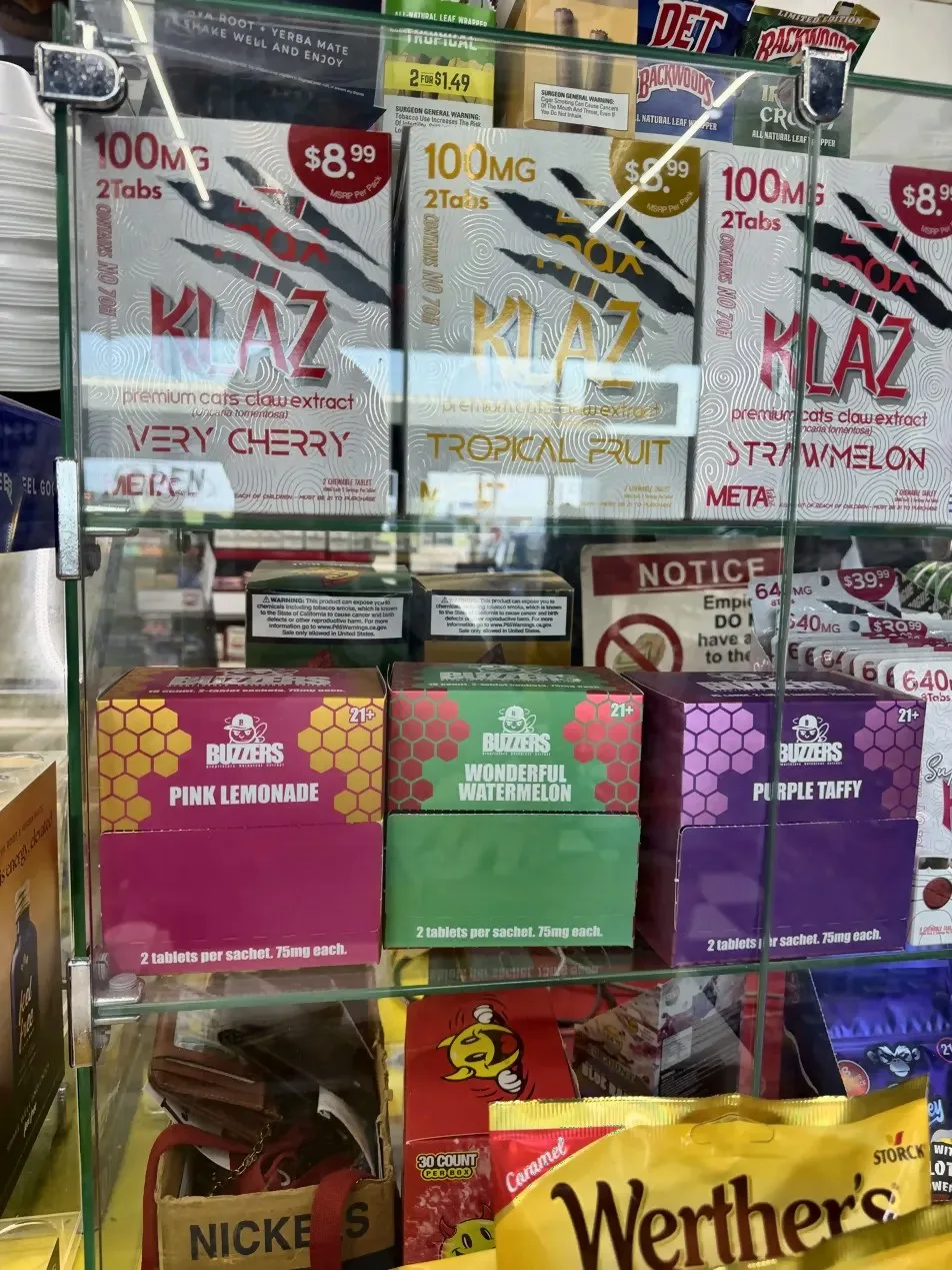 A glass case in a McMinnville, Tennessee store in August 2026: three KLAZ boxes, premium cats claw extract (Uncaria tomentosa), Very Cherry, Tropical Fruit and Strawmelon, 100 mg 2 tabs, with a 7max logo behind the claw marks, the words Contains no 7OH down the side and META at the foot; three Buzzers boxes below.