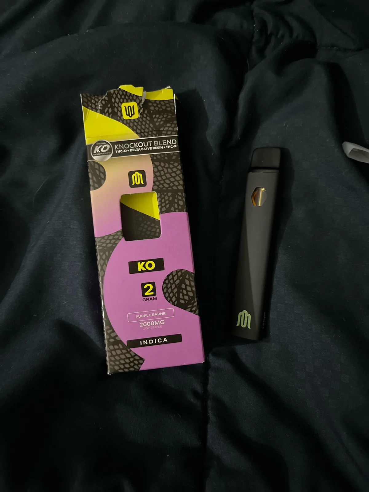 A torn purple and black box printed with snake scales: Knockout Blend, THC-O plus Delta 8 Live Resin plus THC-P, KO, 2 gram, Purple Barnie, 2000 mg disposable, indica, beside a black disposable vape with a green M logo.