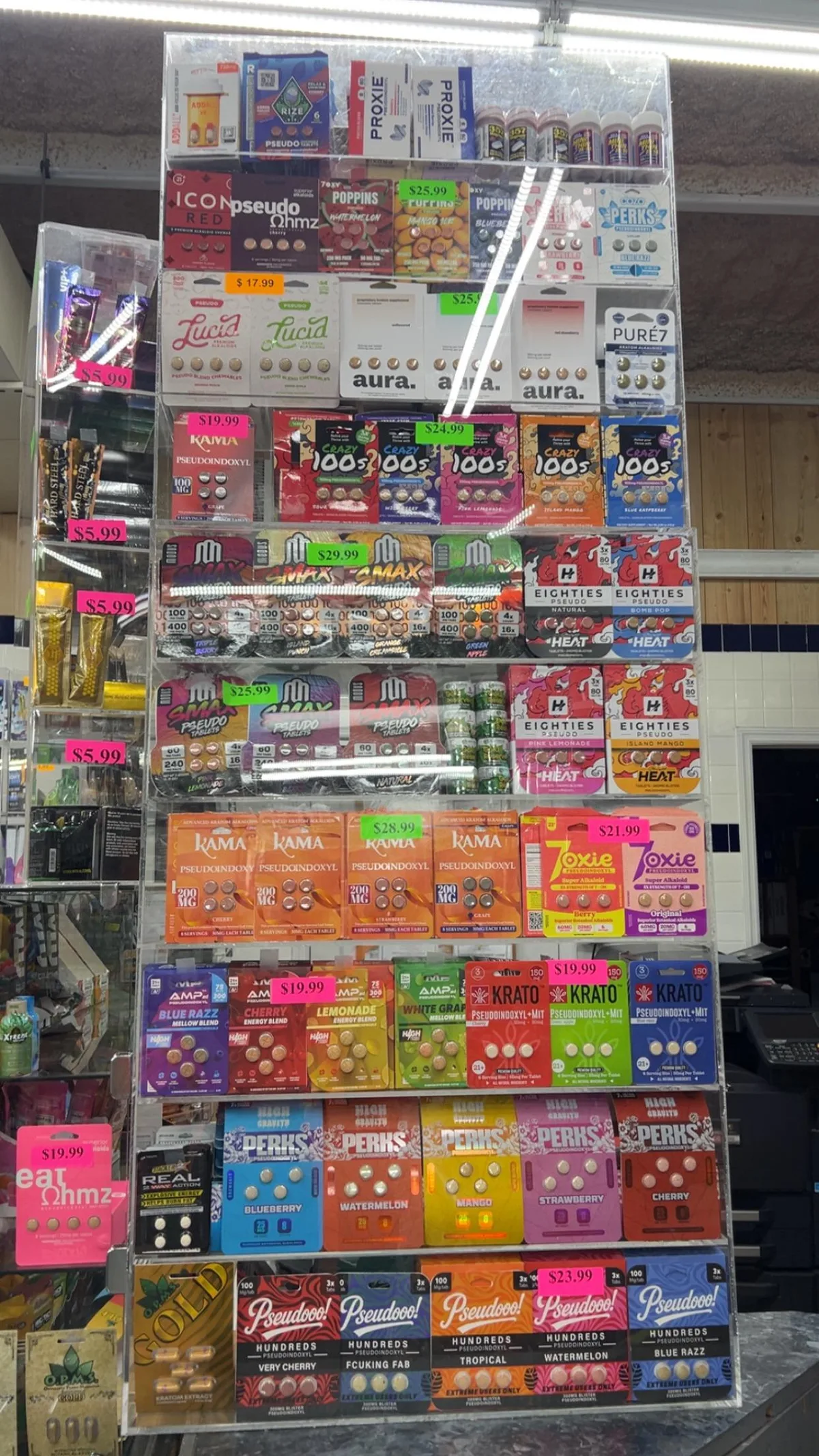 A tall acrylic display in a gas station holding dozens of tablet cards: two rows of Modus Smax packs, 100 mg and 60 mg, among Kama, Crazy 100s, Eighties, Lucid, Aura, Perks, Krato and Pseudooo! cards, with price stickers.