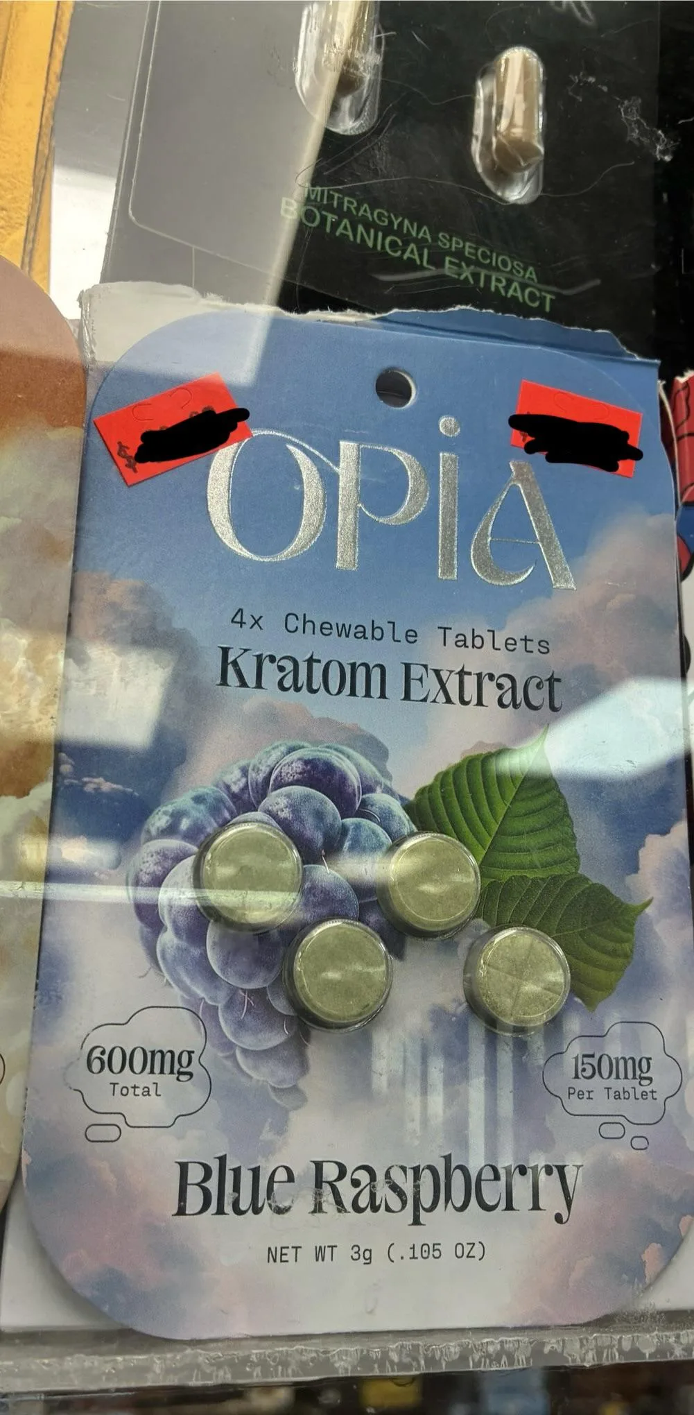 An Opia card on a shop shelf: 4x Chewable Tablets, Kratom Extract, Blue Raspberry, 600mg total, 150mg per tablet, the price stickers blacked out by the poster.