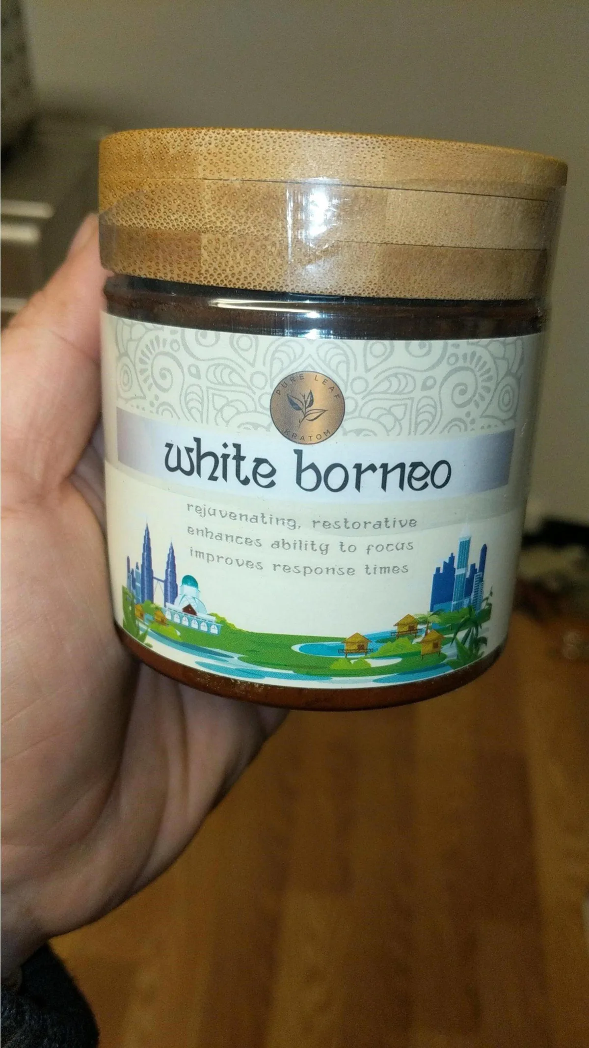 A hand holding a jar with a bamboo lid and a label: the round Pure Leaf Kratom logo, the words white borneo, and the lines rejuvenating, restorative; enhances ability to focus; improves response times, over a drawing of a city skyline and stilt houses.