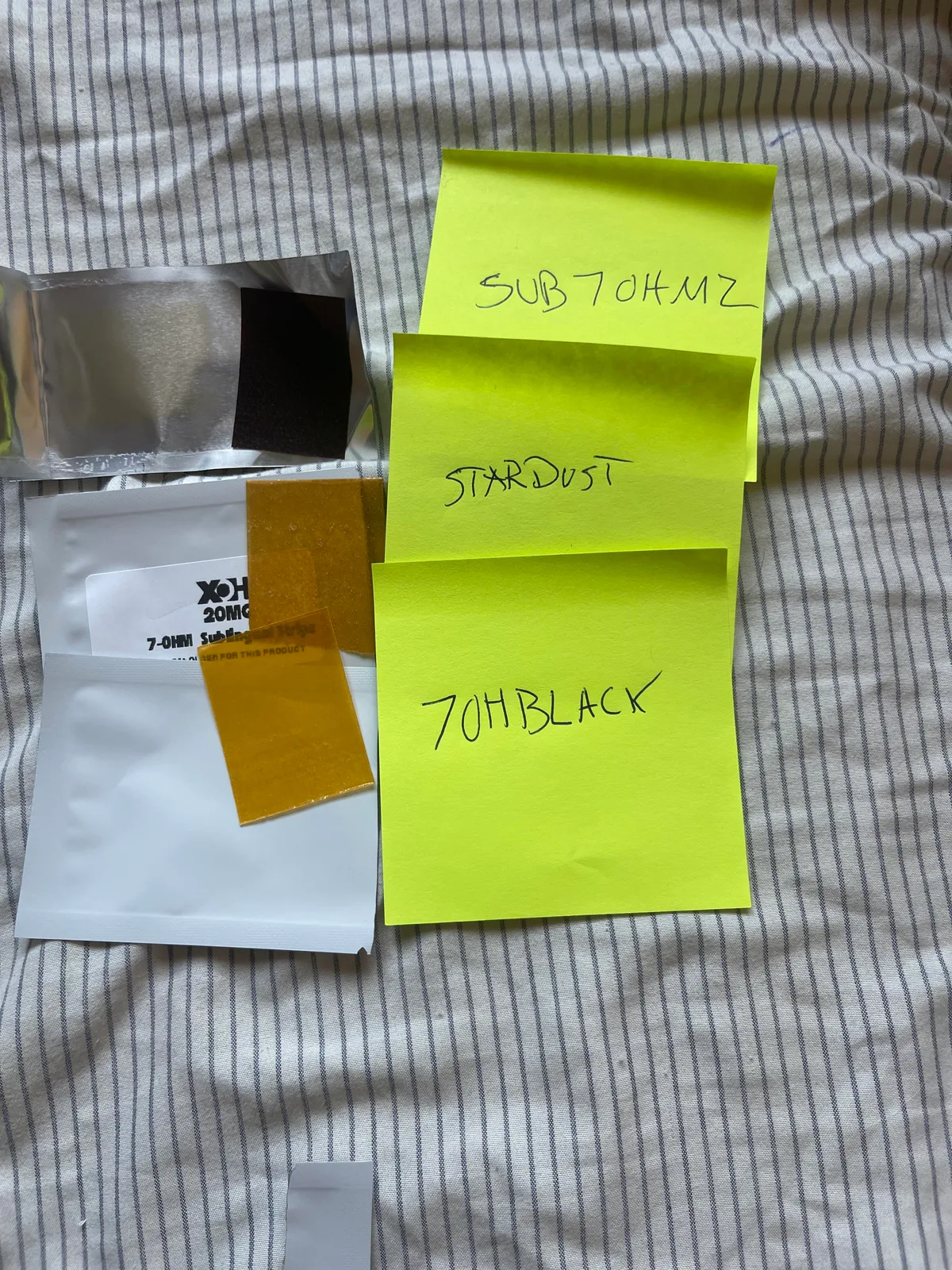 A buyer's photo on a striped sheet: three amber strips on plain white sachets, each under a yellow sticky note reading SUB7OHMZ, STARDUST and 7OHBLACK, with a silver sachet and a printed 20MG 7-OHM sublingual strips label beside them.