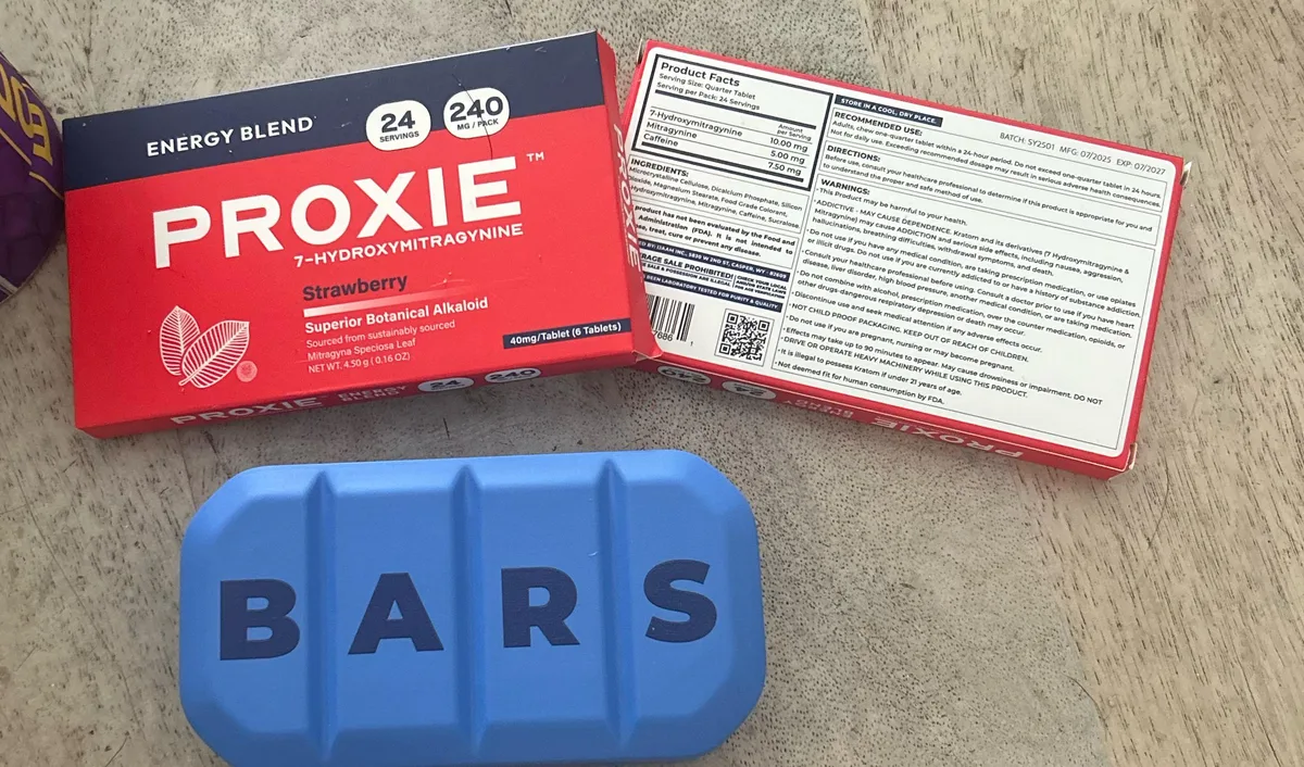 A red Proxie Energy Blend Strawberry box, 24 servings, 240 mg per pack, its back panel beside it, and below them a blue foam object shaped like a scored four-segment pill bar with the letters B A R S across it.