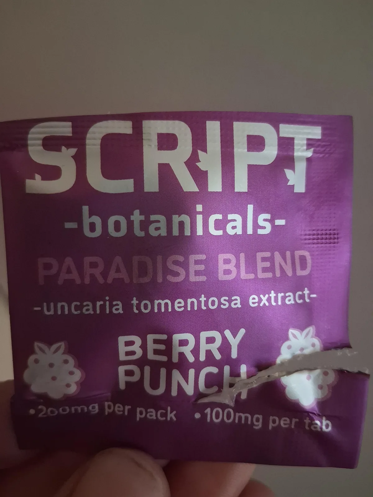 A purple foil sachet held in a hand, printed SCRIPT -botanicals- PARADISE BLEND -uncaria tomentosa extract- BERRY PUNCH, 200mg per pack, 100mg per tab.
