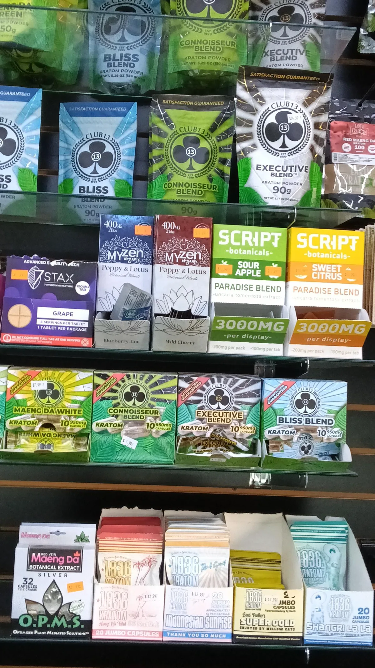 A smoke-shop shelf: kratom powders and capsules, a 7 Stax 7-hydroxymitragynine tablet box, two MyZen Poppy and Lotus boxes, and two Script Botanicals Paradise Blend display boxes in Sour Apple and Sweet Citrus marked 3000MG per display.
