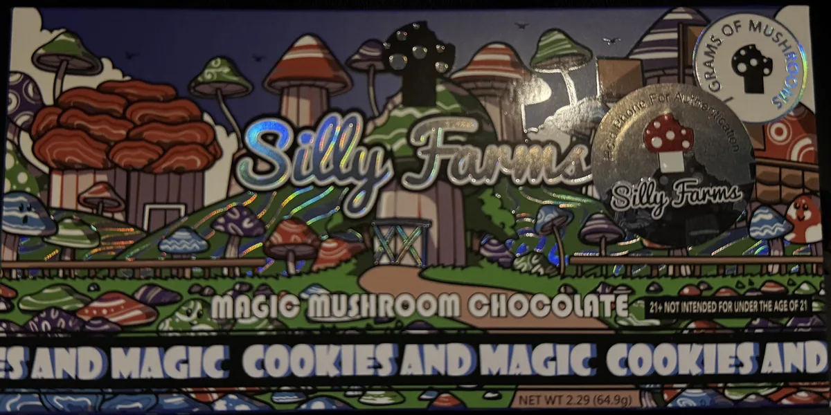 A Cookies and Magic box of Silly Farms Magic Mushroom Chocolate with a holographic sticker reading grams of mushrooms, a round silver sticker reading Hold phone for authentication, and a line reading 21+ not intended for under the age of 21. Net weight 2.29 ounces, 64.9 grams.